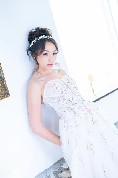 A bride in a white floral wedding dress and headpiece posing against a white wall.