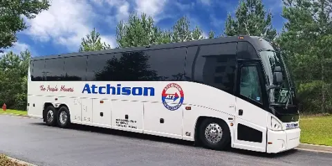 A white and black Atchison Transportation motorcoach parked on a paved road with trees in the background.