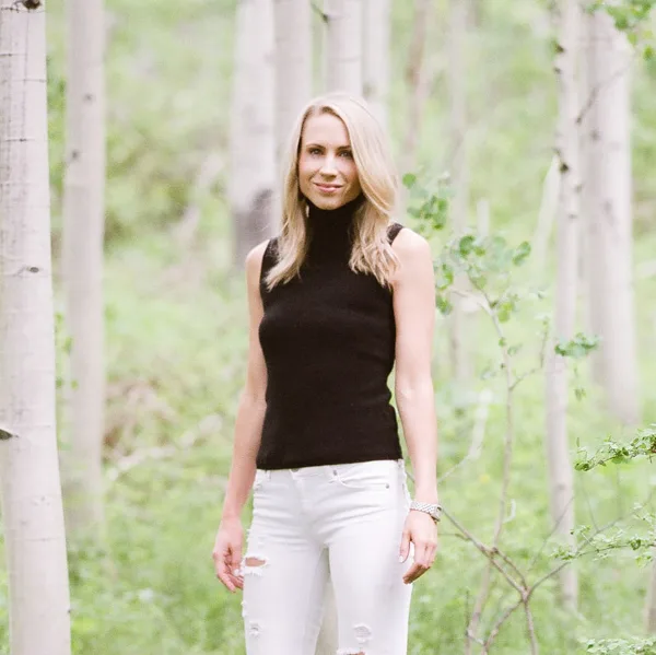Portrait of a blonde woman in a black sleeveless top and white jeans standing in a white birch forest.