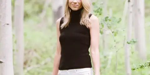 Portrait of a blonde woman in a black sleeveless top and white jeans standing in a white birch forest.