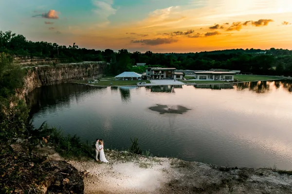 A bride and groom standing on a rocky bluff overlooking a lake and reception hall at sunset.