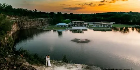 A bride and groom standing on a rocky bluff overlooking a lake and reception hall at sunset.