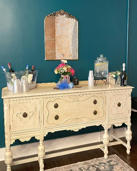A cream-colored vintage sideboard decorated with a floral bouquet, wine bottles, and a beverage dispenser against a teal wall.