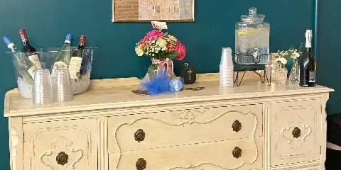 A cream-colored vintage sideboard decorated with a floral bouquet, wine bottles, and a beverage dispenser against a teal wall.