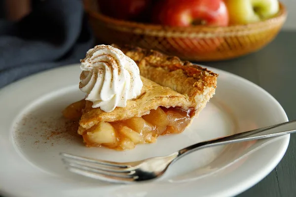 A slice of apple pie topped with a swirl of whipped cream on a white plate with a fork, with a basket of whole apples blurred in the background.