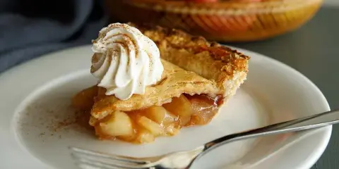A slice of apple pie topped with a swirl of whipped cream on a white plate with a fork, with a basket of whole apples blurred in the background.