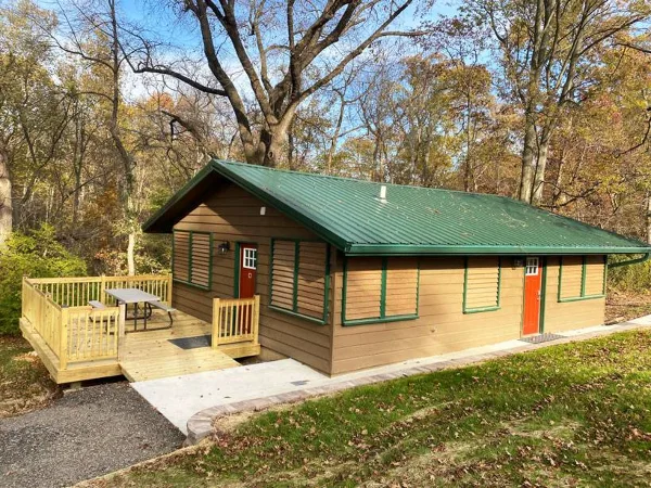 Exterior view of a rustic wooden cottage with a green metal roof and a wooden deck, situated in a wooded area.