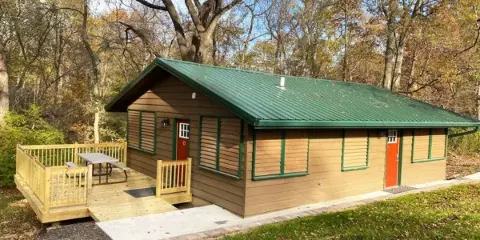 Exterior view of a rustic wooden cottage with a green metal roof and a wooden deck, situated in a wooded area.