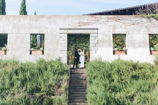 Bride and groom kissing on stone steps in front of a concrete building with greenery