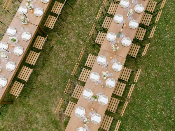 Overhead view of an outdoor wedding reception setup with long wooden tables, white place settings, and floral centerpieces on a grassy lawn.
