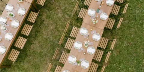 Overhead view of an outdoor wedding reception setup with long wooden tables, white place settings, and floral centerpieces on a grassy lawn.
