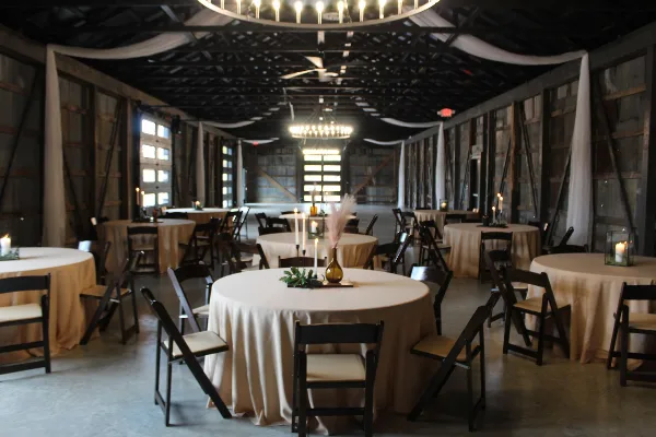 Interior view of a rustic event venue with round tables, chairs, and chandeliers.