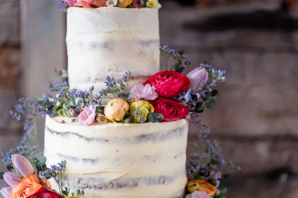 A two-tier semi-naked wedding cake decorated with red, pink, and yellow fresh flowers and greenery.