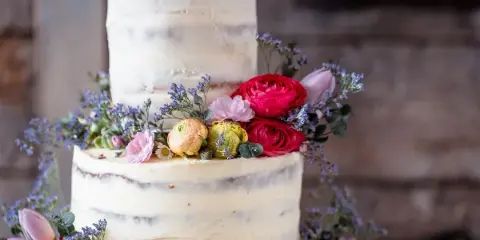 A two-tier semi-naked wedding cake decorated with red, pink, and yellow fresh flowers and greenery.