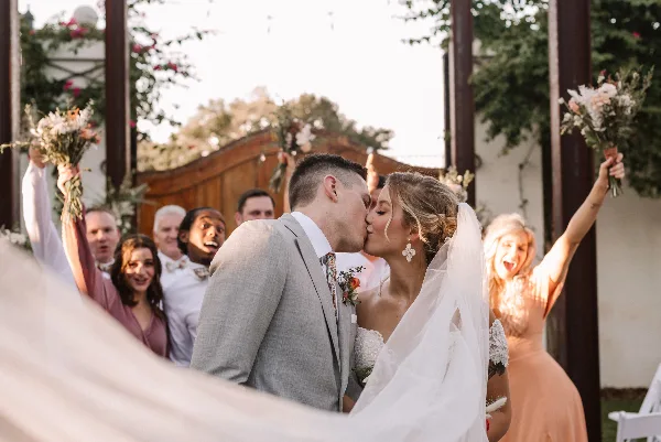 A bride and groom kissing during a wedding ceremony with guests cheering in the background.
