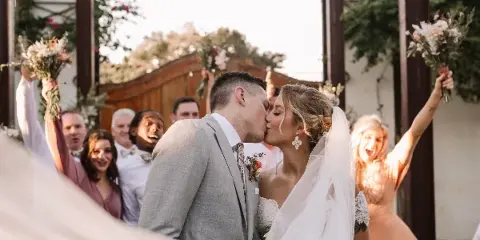 A bride and groom kissing during a wedding ceremony with guests cheering in the background.