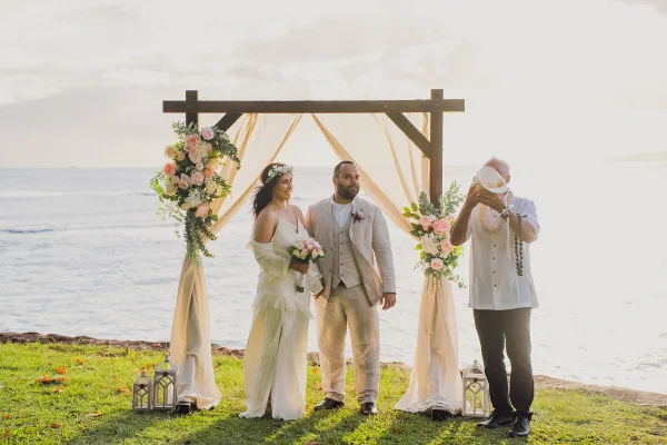 A bride and groom standing under a floral arch on a beach with an officiant during a wedding ceremony.