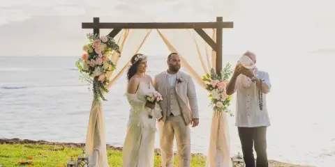 A bride and groom standing under a floral arch on a beach with an officiant during a wedding ceremony.