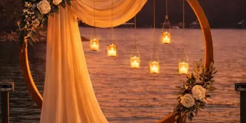 A wooden wedding arch decorated with white roses and hanging candles on a wooden dock overlooking a lake at sunset.