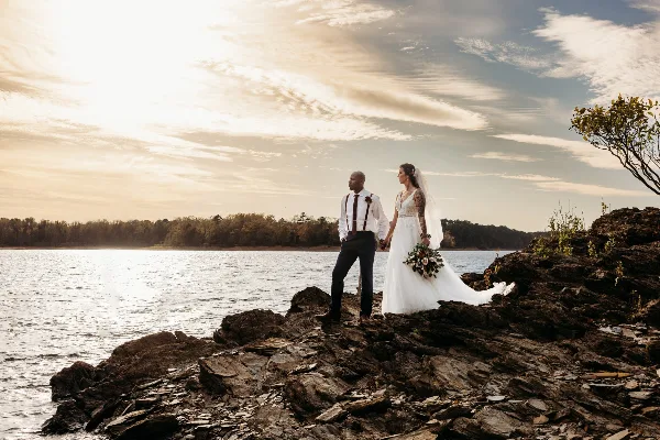 A bride and groom standing on a rocky shore by a lake during a wedding.