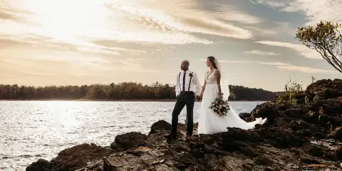 A bride and groom standing on a rocky shore by a lake during a wedding.