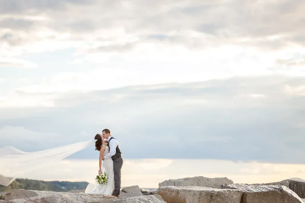 A bride and groom kissing on a rocky shoreline with a flowing veil against a cloudy sky.