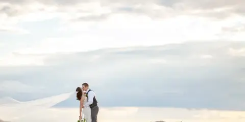 A bride and groom kissing on a rocky shoreline with a flowing veil against a cloudy sky.