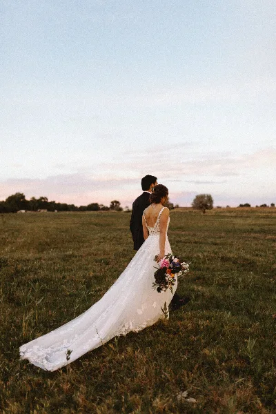 A bride and groom standing in a grassy field at sunset, with the bride holding a bouquet.