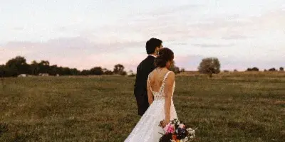 A bride and groom standing in a grassy field at sunset, with the bride holding a bouquet.