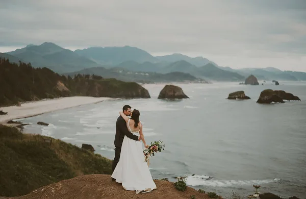 A bride and groom embracing on a coastal cliff overlooking the ocean and mountains.