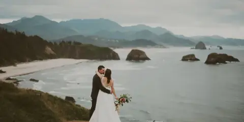 A bride and groom embracing on a coastal cliff overlooking the ocean and mountains.