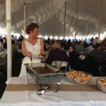 A woman in a white dress serving food from a chafing dish at a large event under a white tent.