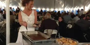 A woman in a white dress serving food from a chafing dish at a large event under a white tent.
