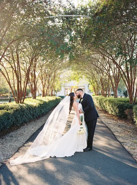A bride and groom kissing on a tree-lined path during a wedding.