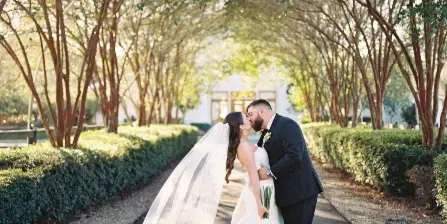 A bride and groom kissing on a tree-lined path during a wedding.