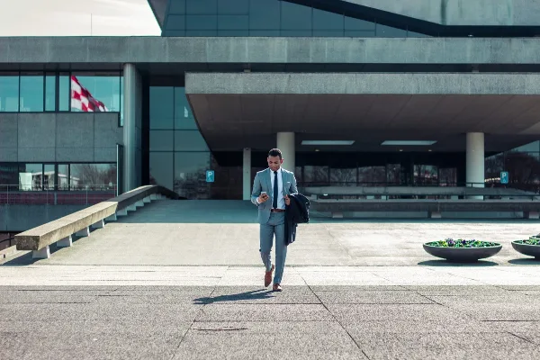 A man in a light blue suit walks while checking his phone in front of a modern building.