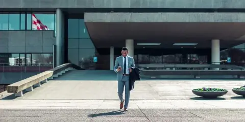 A man in a light blue suit walks while checking his phone in front of a modern building.