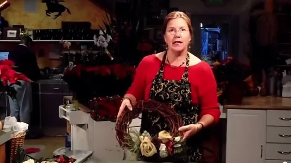 A woman in a red shirt and apron stands in a flower shop holding a floral wreath arrangement with roses and greenery.