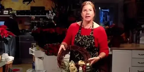 A woman in a red shirt and apron stands in a flower shop holding a floral wreath arrangement with roses and greenery.