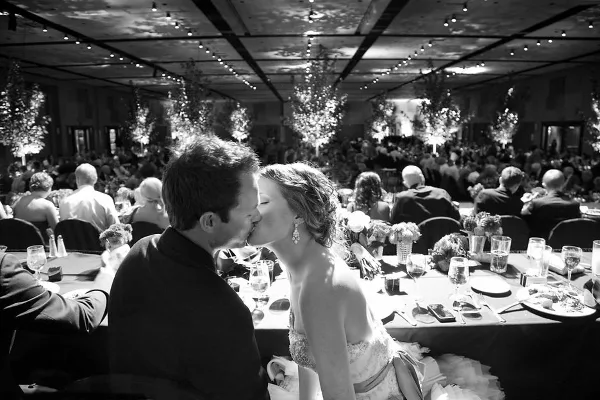 A black and white photograph of a bride and groom kissing at their wedding reception table, with guests and floral decorations in the background.