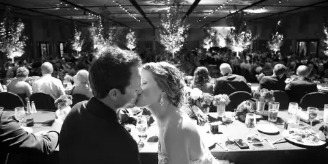 A black and white photograph of a bride and groom kissing at their wedding reception table, with guests and floral decorations in the background.