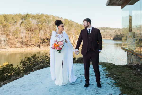 A bride in a white gown and fur stole holding hands with a groom in a maroon suit, standing on a snowy patch of ground near a lake and modern glass building.