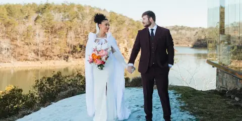 A bride in a white gown and fur stole holding hands with a groom in a maroon suit, standing on a snowy patch of ground near a lake and modern glass building.