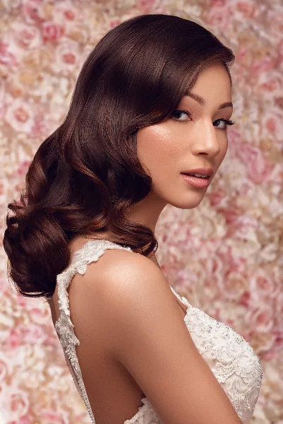 A bride in a white lace wedding dress with styled wavy hair and professional makeup, posing against a floral background.