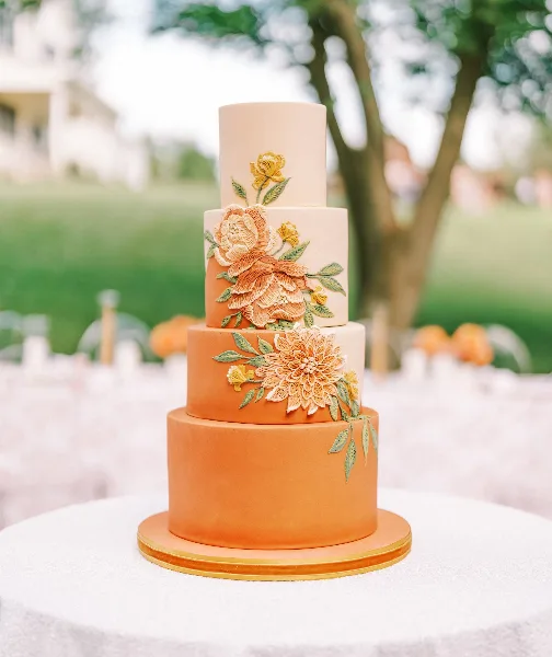 A four-tiered wedding cake with smooth orange and cream fondant, decorated with intricate hand-piped sugar flowers in shades of peach and yellow, displayed on a white table outdoors.