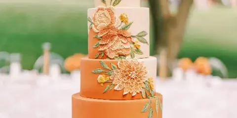 A four-tiered wedding cake with smooth orange and cream fondant, decorated with intricate hand-piped sugar flowers in shades of peach and yellow, displayed on a white table outdoors.