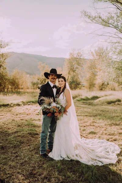 A bride and groom standing together in a grassy field with mountains in the background, holding a bouquet.