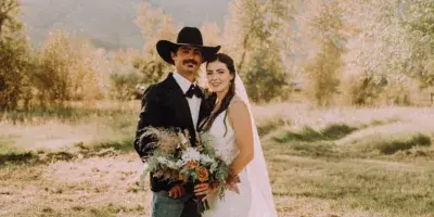 A bride and groom standing together in a grassy field with mountains in the background, holding a bouquet.