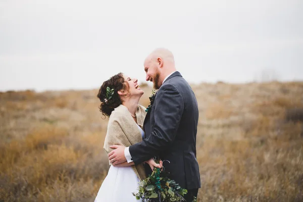 A bride and groom laughing together in a field of dry grass, with the bride holding a bouquet.