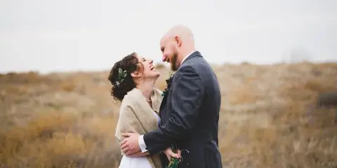 A bride and groom laughing together in a field of dry grass, with the bride holding a bouquet.